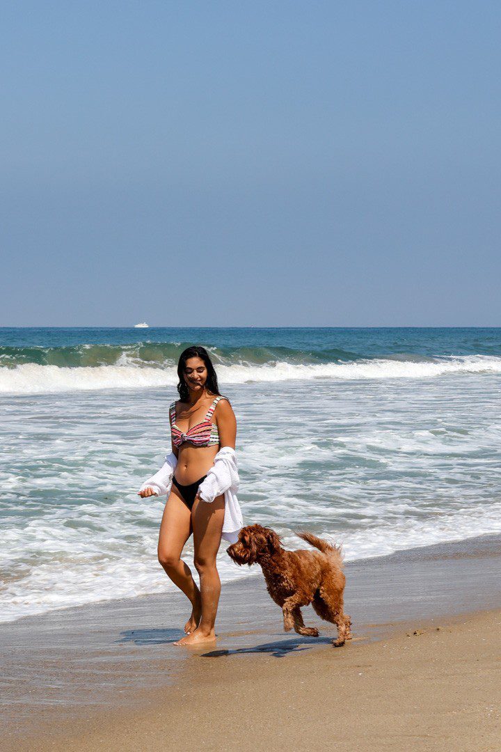 A woman in a bikini walking her dog on the beach.