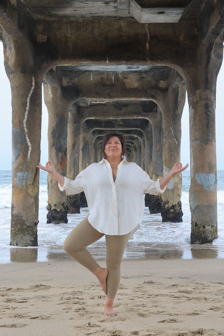 A woman doing yoga on the beach under a pier.