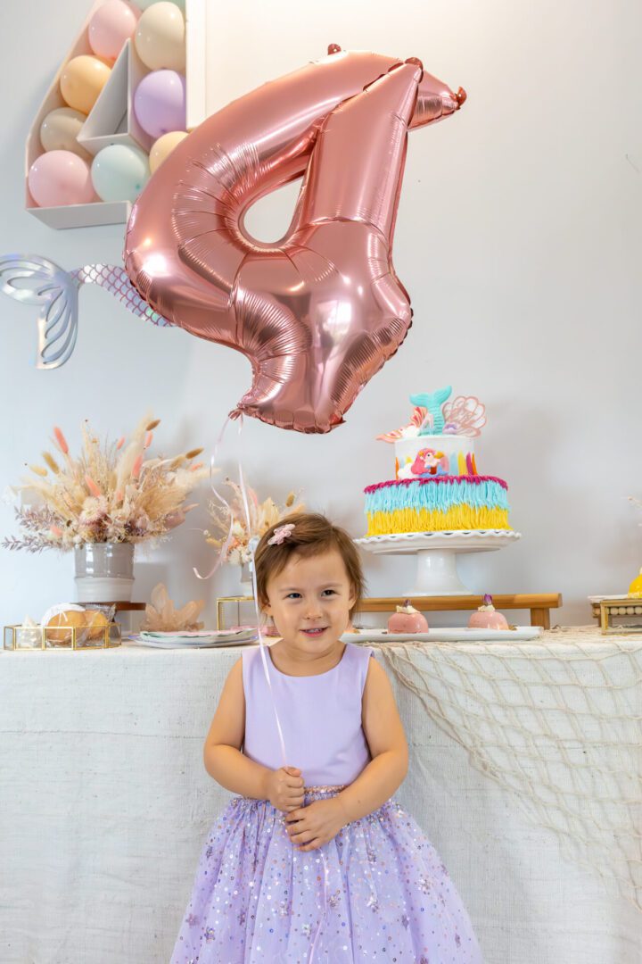 A little girl in a purple dress standing in front of a table with balloons.