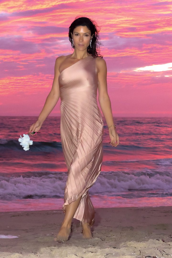 A woman in a pink dress walking on the beach at sunset.